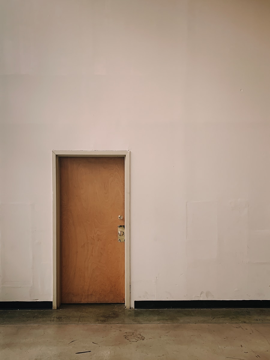 Drzwi z drewna na tle białej ściany, z widoczną podłogą, w minimalistycznym wnętrzu.
