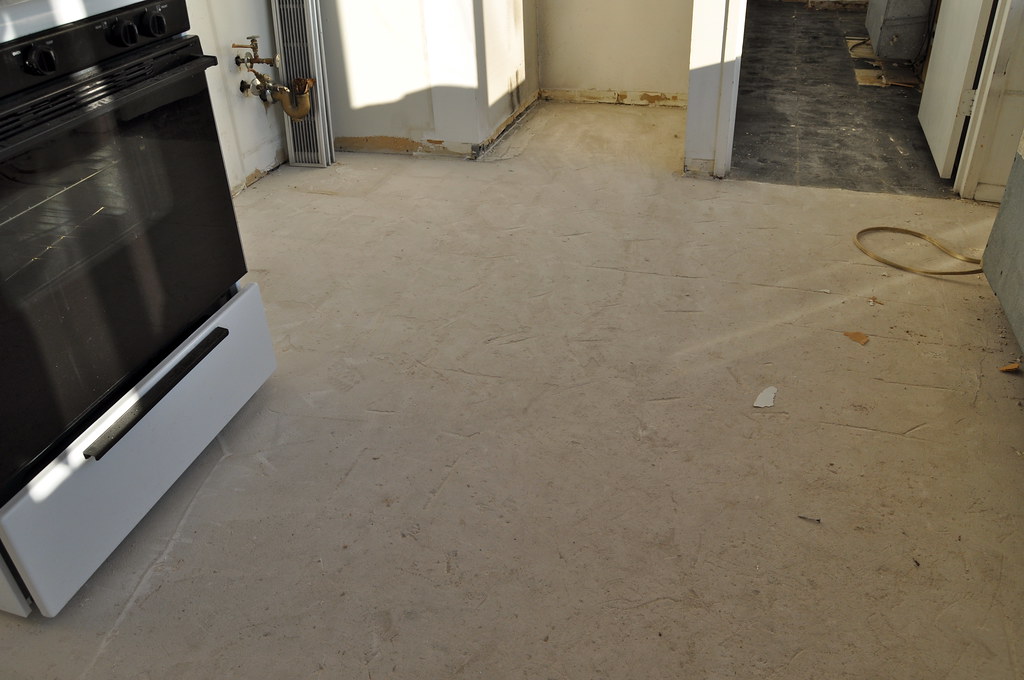 Kuchnia w trakcie remontu, widoczny czarny piekarnik i jasna podłoga. W tle fragmenty ścian oraz węże instalacyjne.